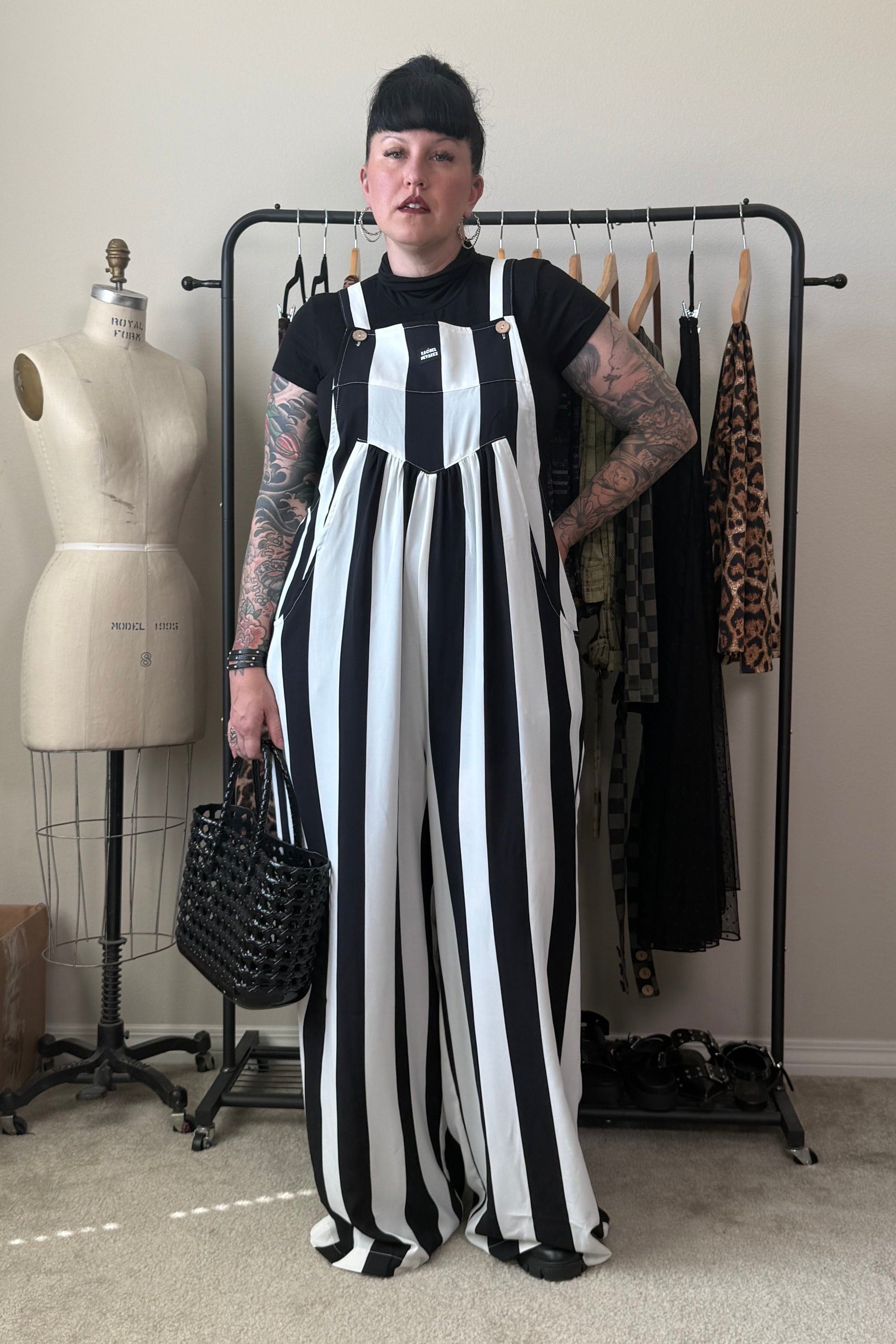A person stands in a room wearing a black and white vertically striped jumpsuit over a black t-shirt, holding a black woven handbag. A dress form, clothing rack, and room elements are visible in the background.
