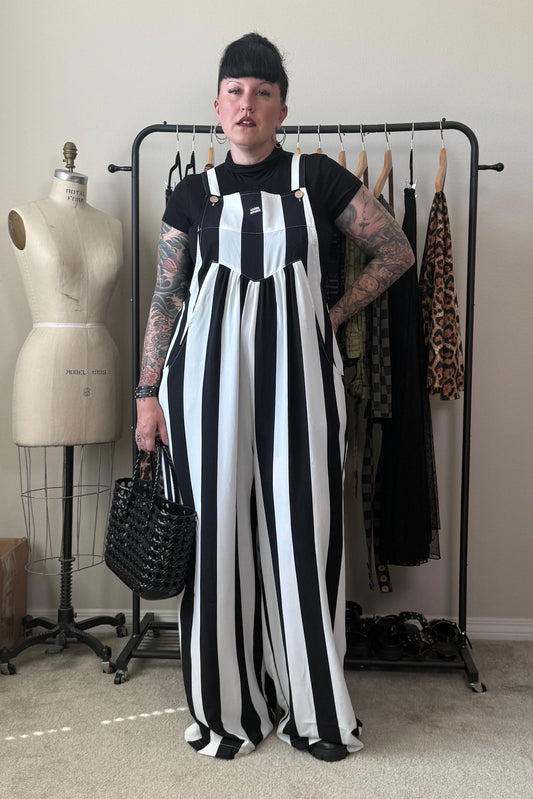 A person stands in a room wearing a black and white vertically striped jumpsuit over a black t-shirt, holding a black woven handbag. A dress form, clothing rack, and room elements are visible in the background.