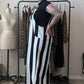 A woman with short black hair and tattoos stands in a room wearing a sleeveless black and white vertically striped maxi dress. Behind her is a clothing rack with patterned dresses and a dress form mannequin against a plain beige wall.