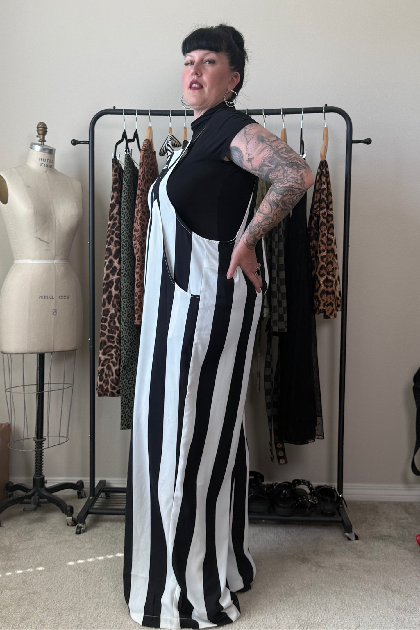 A woman with short black hair and tattoos stands in a room wearing a sleeveless black and white vertically striped maxi dress. Behind her is a clothing rack with patterned dresses and a dress form mannequin against a plain beige wall.