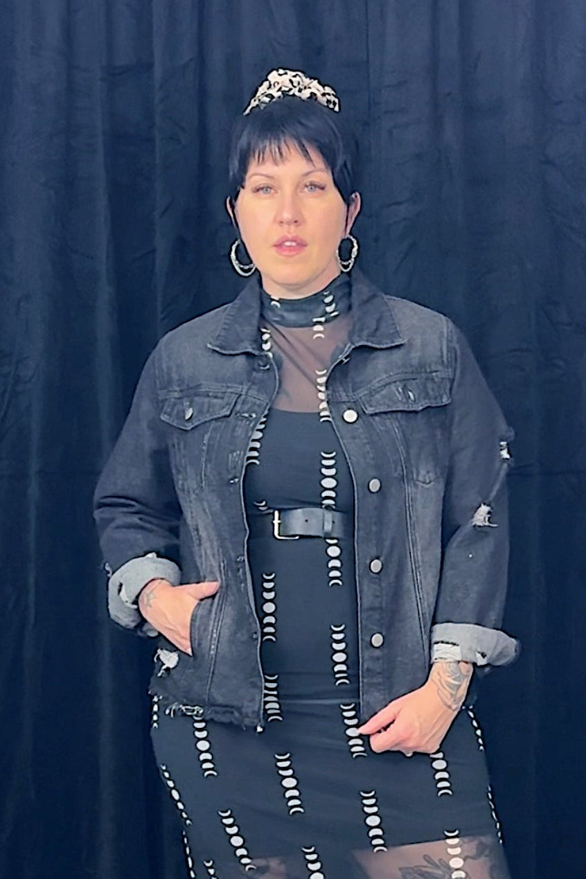 Person wearing a denim jacket and black dress with white patterns against a dark curtain background