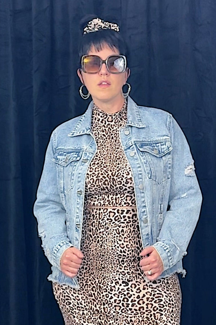 Person wearing a leopard print dress and denim jacket against a dark blue curtain.