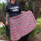 Person wearing a black t-shirt and pink skirt with green patterns, standing outdoors with palm trees in the background.