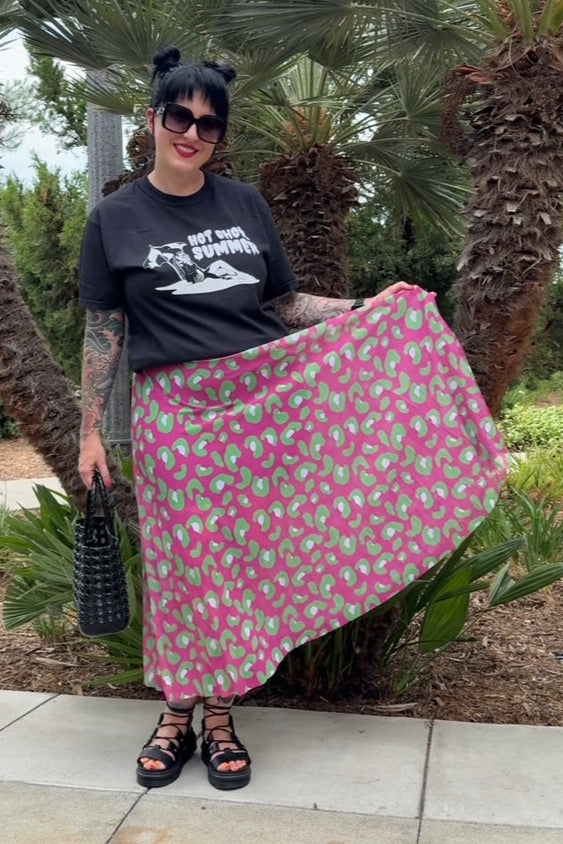 Person wearing a black t-shirt and pink skirt with green patterns, standing outdoors with palm trees in the background.