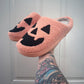 Pink slippers with black jack-o'-lantern faces on a white paneled wall background.