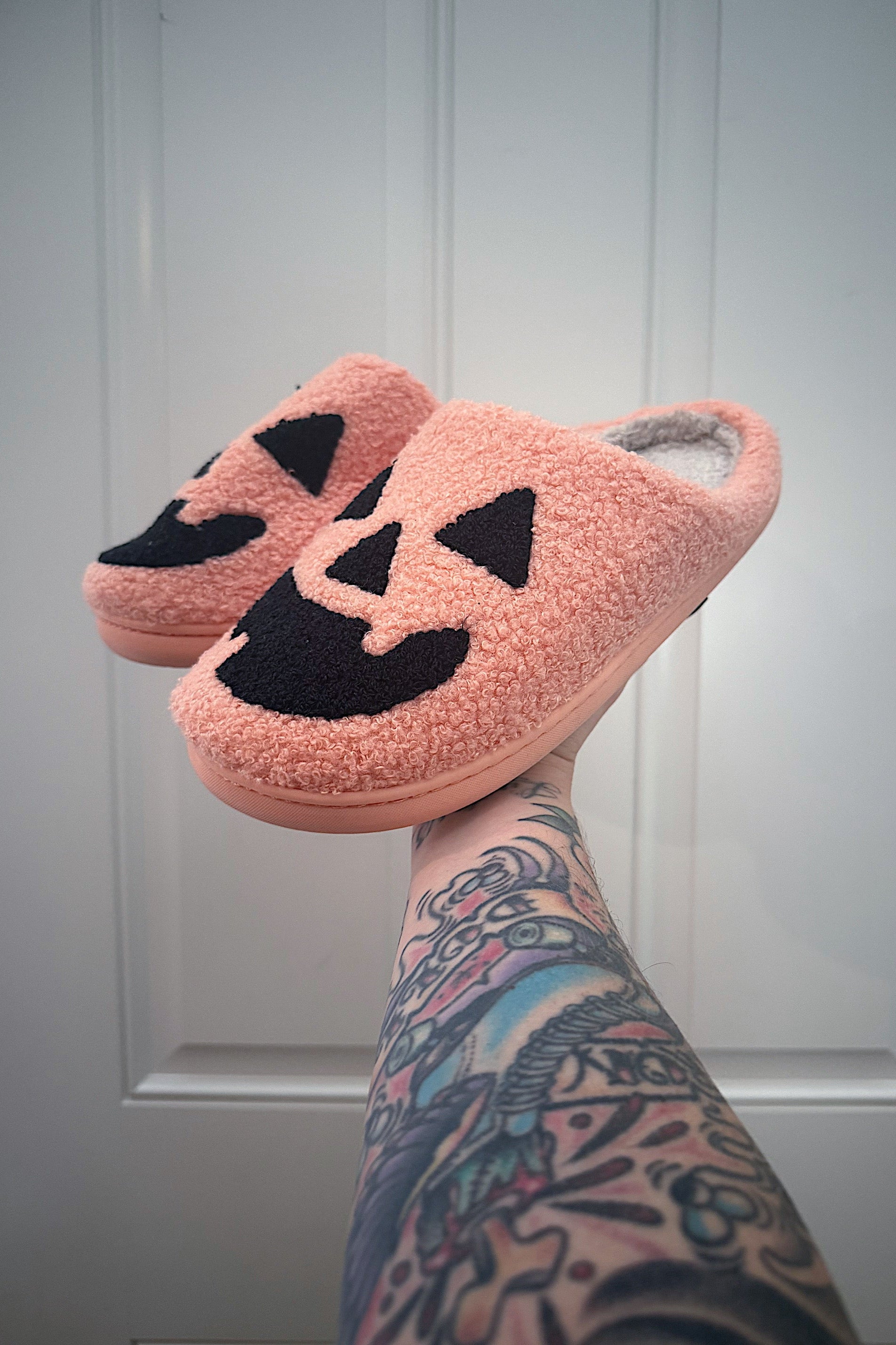 Pink slippers with black jack-o'-lantern faces on a white paneled wall background.