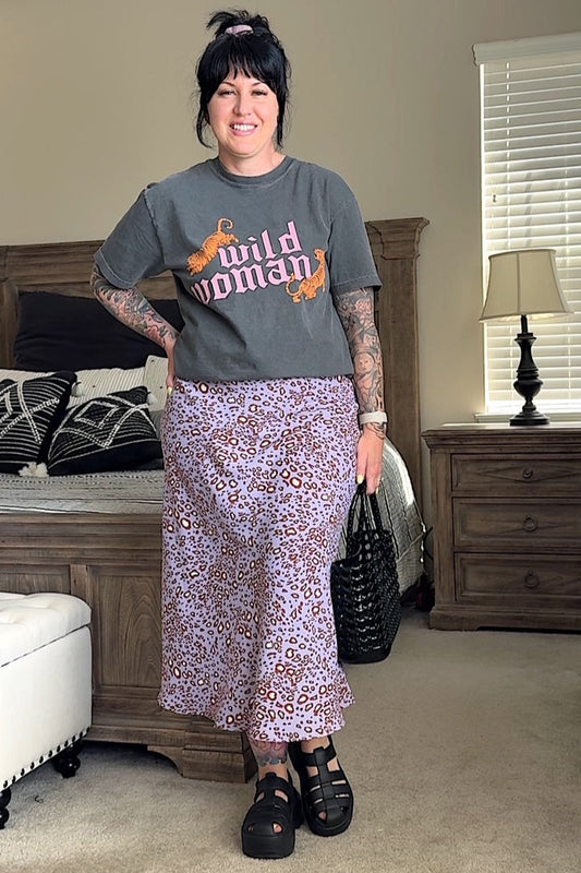 A person wearing a gray t-shirt with the text 'Wild Woman' and a midi-length A-line skirt with a lavender leopard print. Accessories include black strappy sandals and a black bag.
