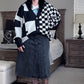 Woman standing in a bedroom wearing a black and white checkered cardigan, denim skirt, and black boots.