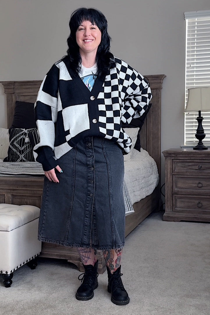 Woman standing in a bedroom wearing a black and white checkered cardigan, denim skirt, and black boots.