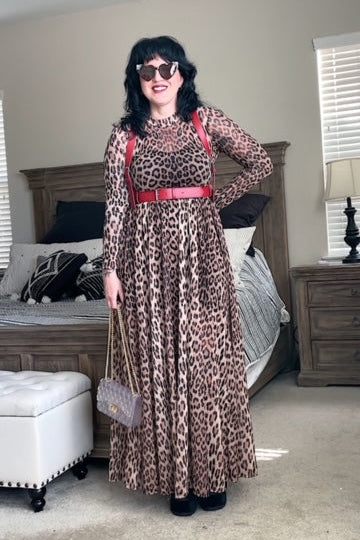 A person wearing a leopard print dress with a red vegan leather fashion harness, standing in a room with a white chair and wooden furniture in the background.