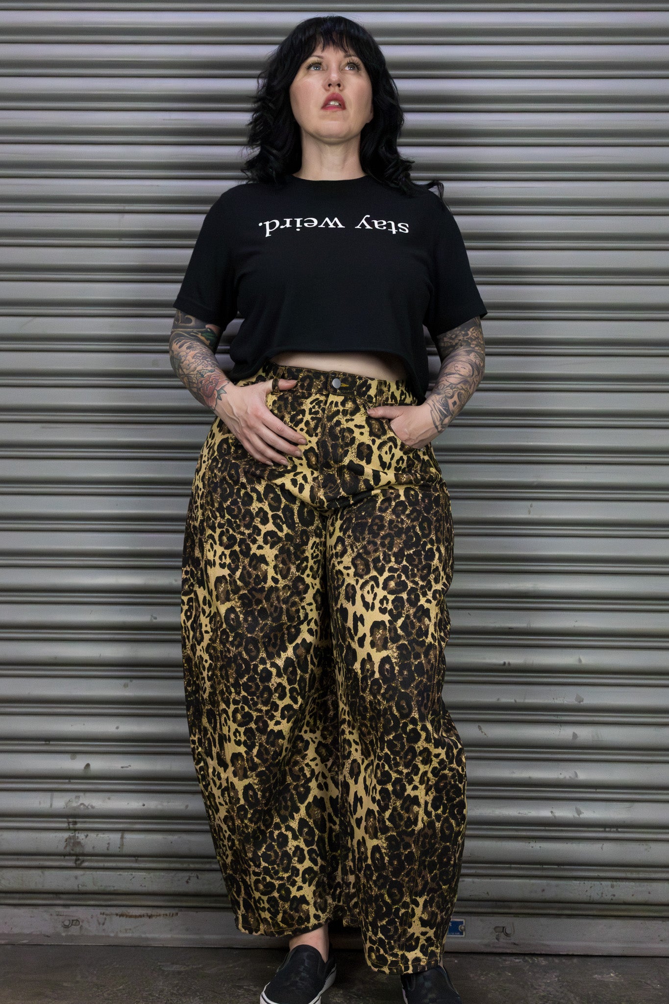 Person wearing a black t-shirt with text and leopard print pants standing in front of a metal shutter.