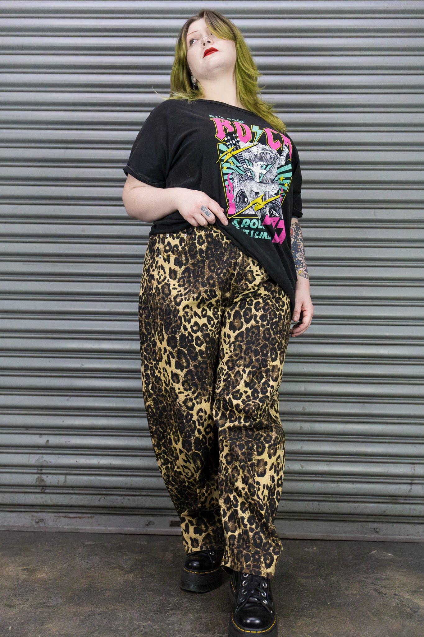 Person wearing a black graphic t-shirt and leopard print pants standing in front of a metal shutter.