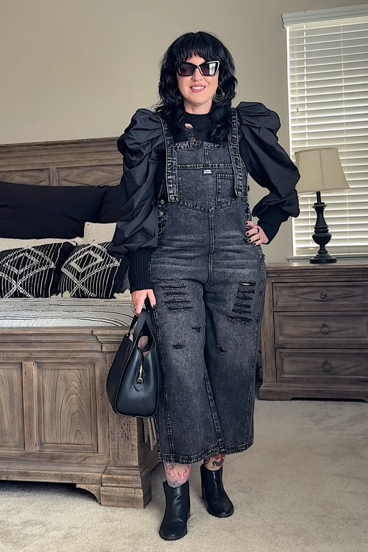 Person wearing a black denim overalls and black top in a bedroom setting.