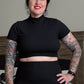 Person wearing a black turtleneck and black skirt with tattoos on arms, standing in a bedroom.