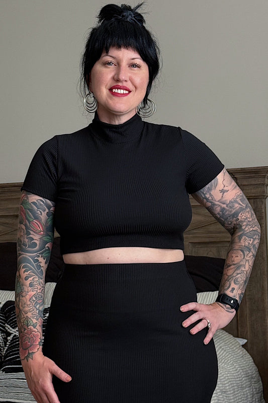 Person wearing a black turtleneck and black skirt with tattoos on arms, standing in a bedroom.