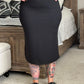 Person wearing a black dress and high-heeled shoes with tattoos on their arms and legs, standing in a room.