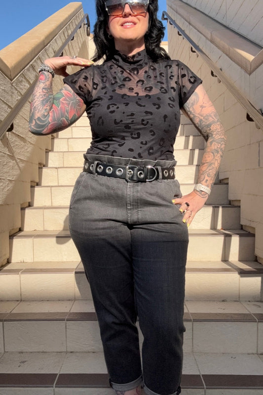 A person wearing a black mesh short sleeve top with a leopard print design, paired with dark jeans and a belt, standing on steps.