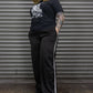 Person wearing a black t-shirt with a graphic design and black pants with white stripes, standing against a metallic shutter.