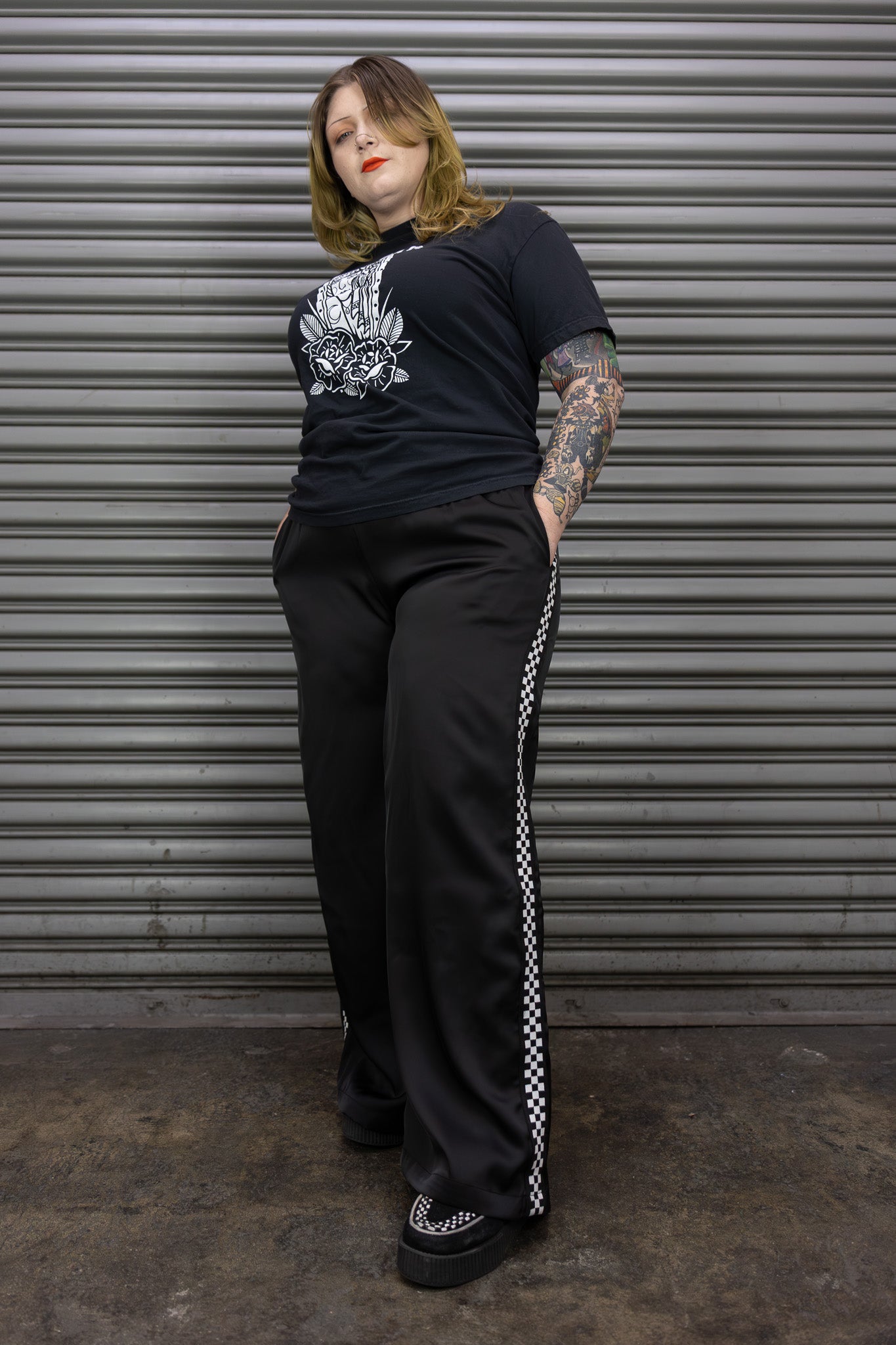 Person wearing a black t-shirt with a graphic design and black pants with white stripes, standing against a metallic shutter.