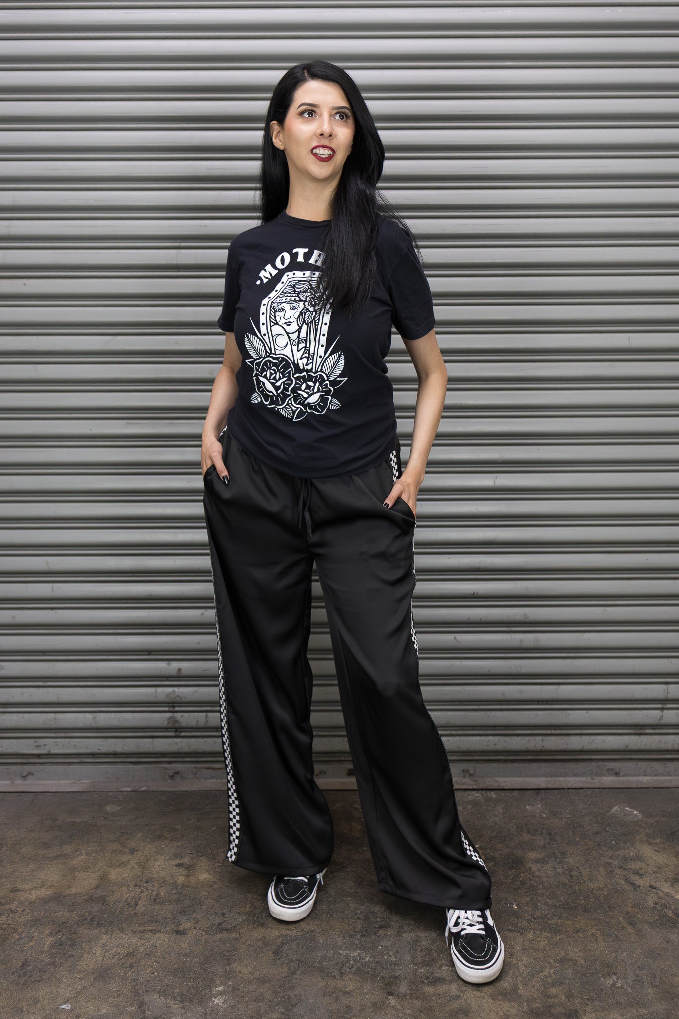 Person wearing a black t-shirt with graphic design and black pants standing in front of a metallic shutter.