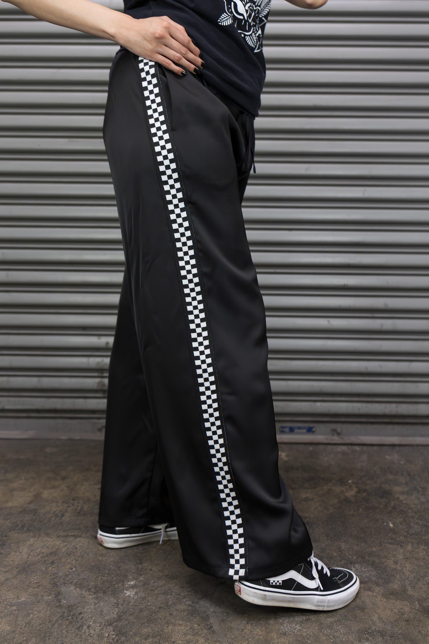 Person wearing black pants with white checkered stripe, standing against a metallic shutter background.