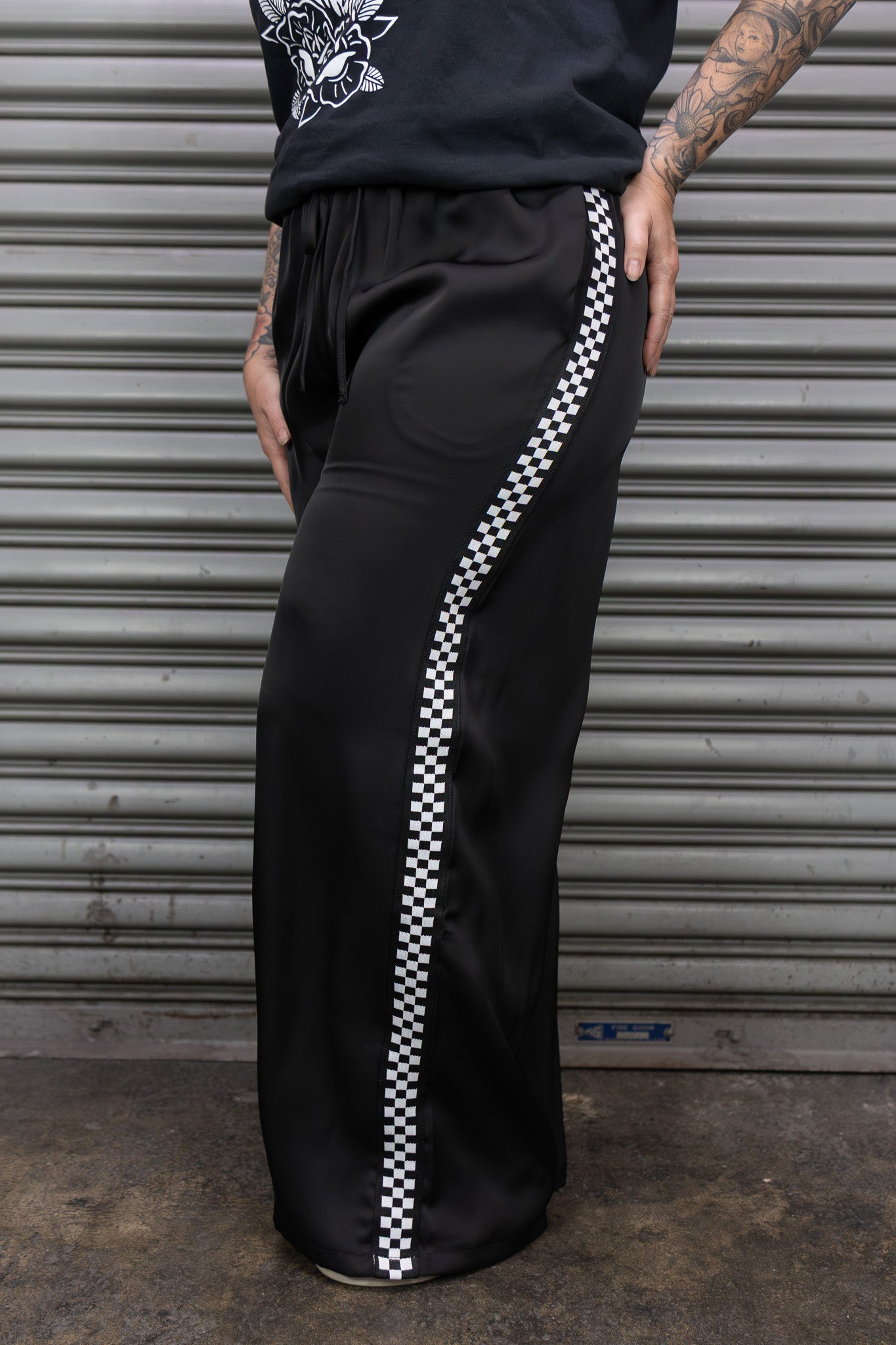 Person wearing black pants with a white checkered stripe in front of a gray metal shutter.