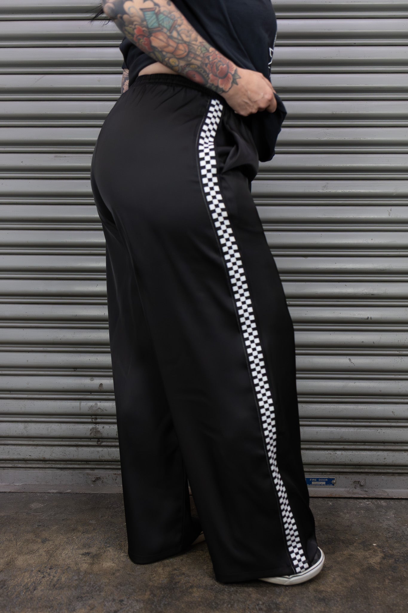 Person wearing black wide-leg pants with white checkered stripe against a metal shutter background.