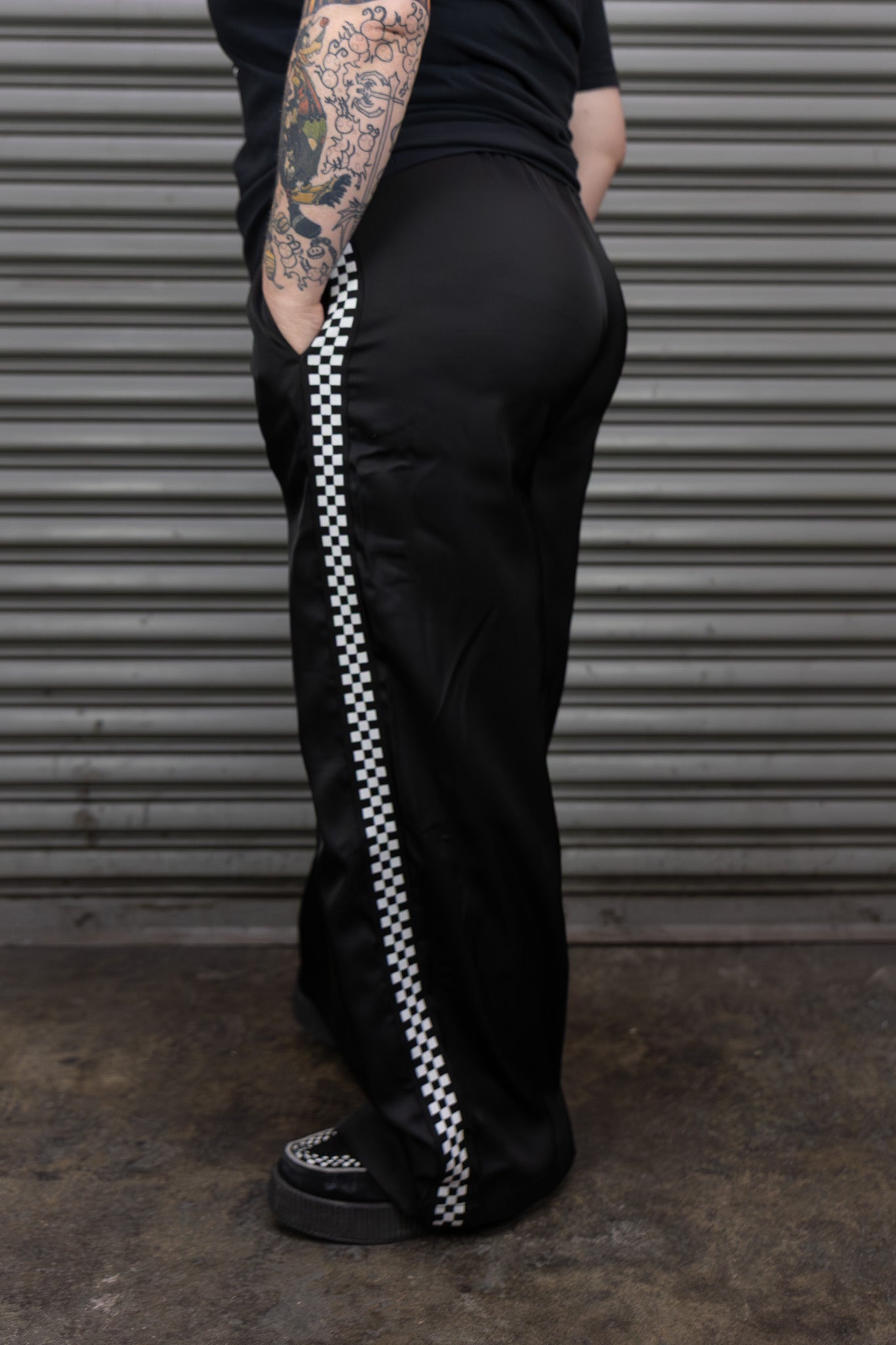 Person wearing black pants with a white checkered stripe, standing against a metallic shutter.