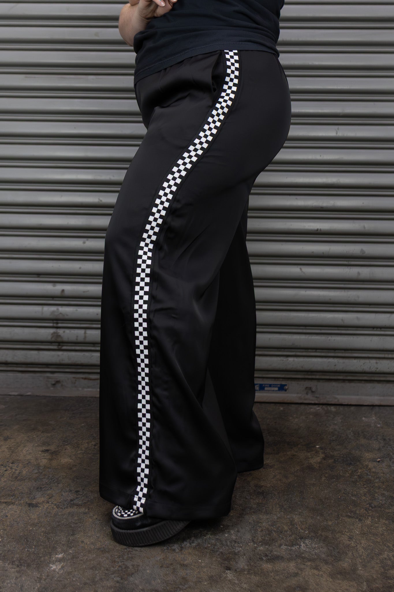 Person wearing black pants with a white checkered stripe against a metallic shutter background
