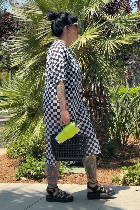 Person wearing a black and white checkered dress with tattoos, standing outdoors near greenery.