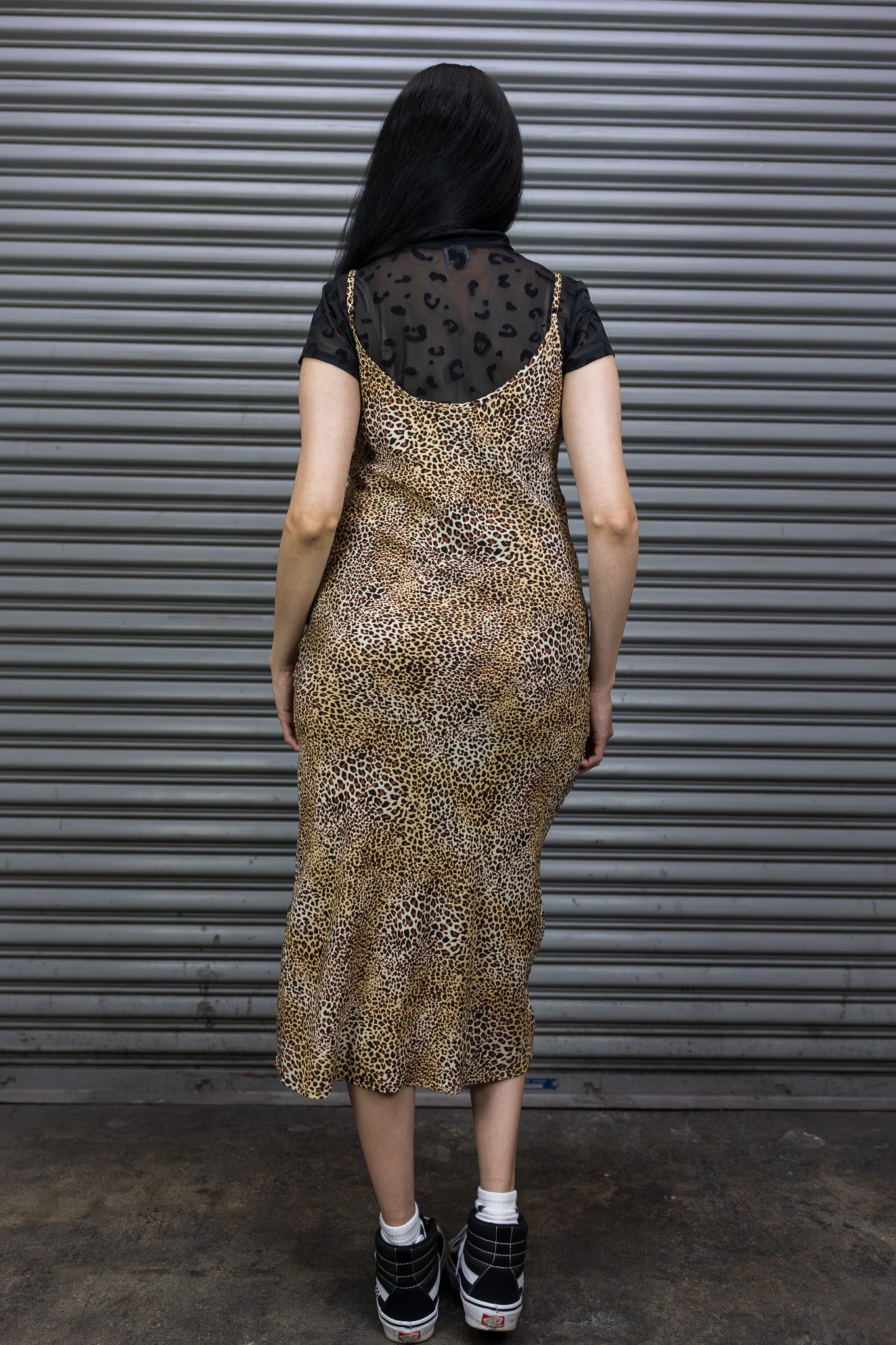 Person wearing a leopard print dress over a black top with sheer details, standing against a metallic shutter.