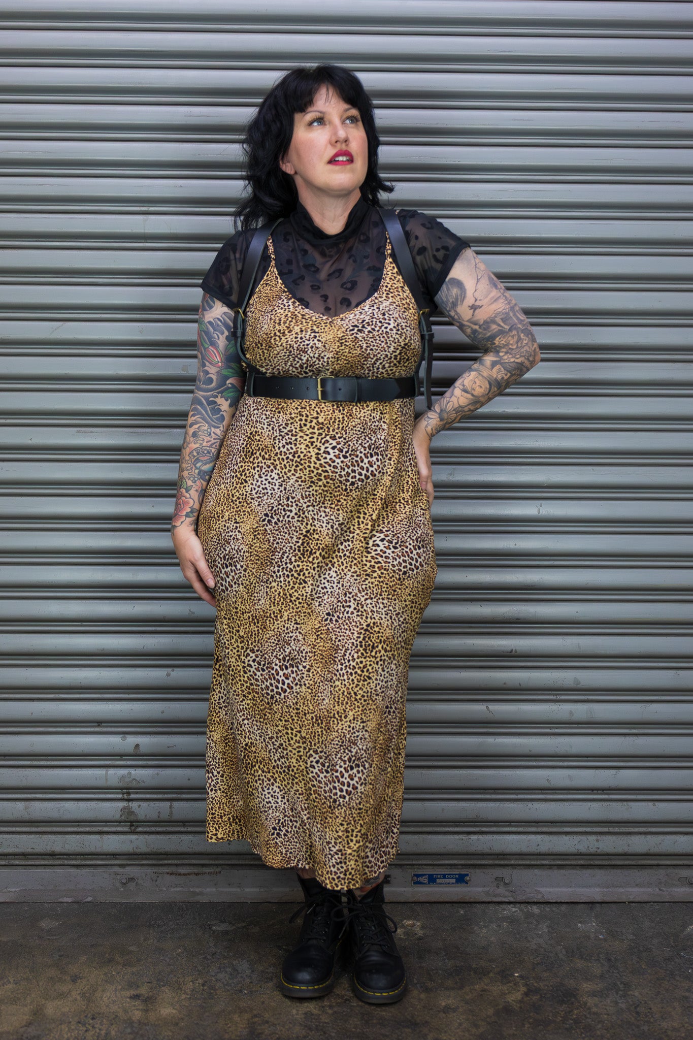 Person wearing a patterned dress with tattoos standing in front of a metal shutter.