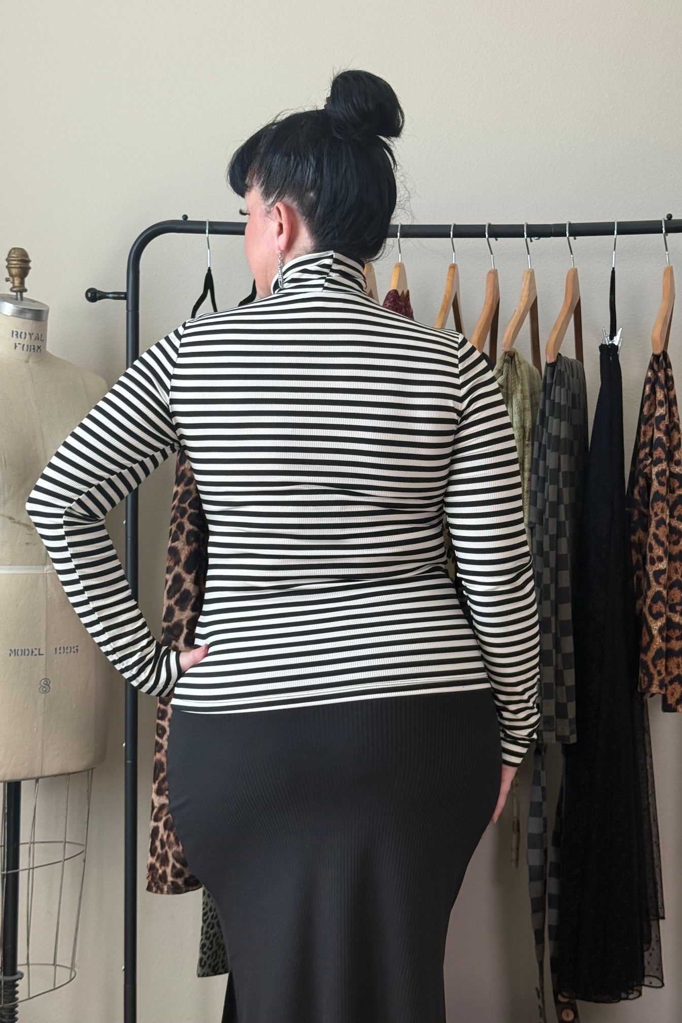 A woman viewed from behind wearing a black and white horizontally striped mock neck top and a black skirt, standing in front of a clothing rack.