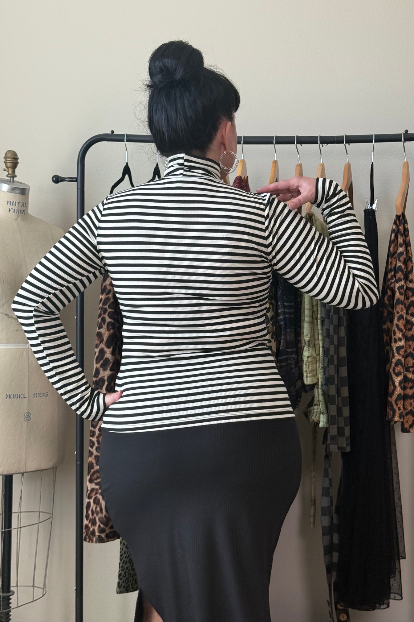 A woman viewed from behind wearing a black and white striped long-sleeve mock neck top and a black skirt, standing in front of a clothing rack.