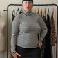 A woman stands indoors wearing a fitted black-and-white horizontally striped mock-neck long-sleeve top with a black skirt. She is posed in front of a clothing rack with other garments hanging on wooden hangers, with a dress form visible to the left.