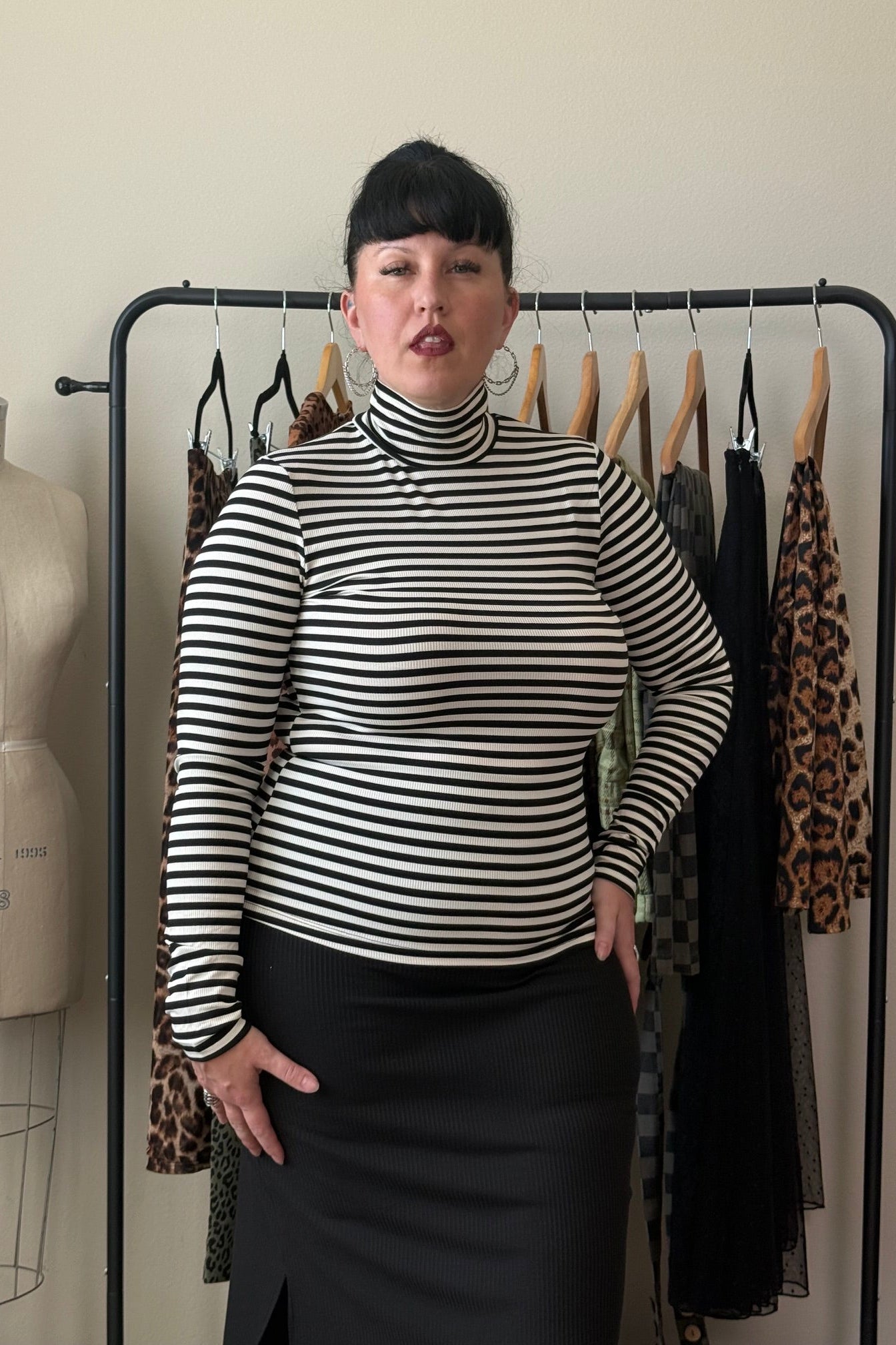A woman stands indoors wearing a fitted black-and-white horizontally striped mock-neck long-sleeve top with a black skirt. She is posed in front of a clothing rack with other garments hanging on wooden hangers, with a dress form visible to the left.