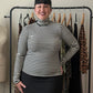 A woman with short black hair stands smiling in front of a clothing rack, wearing a black and white horizontally striped mock neck long-sleeve top and a black skirt. A dress form is visible to her left.