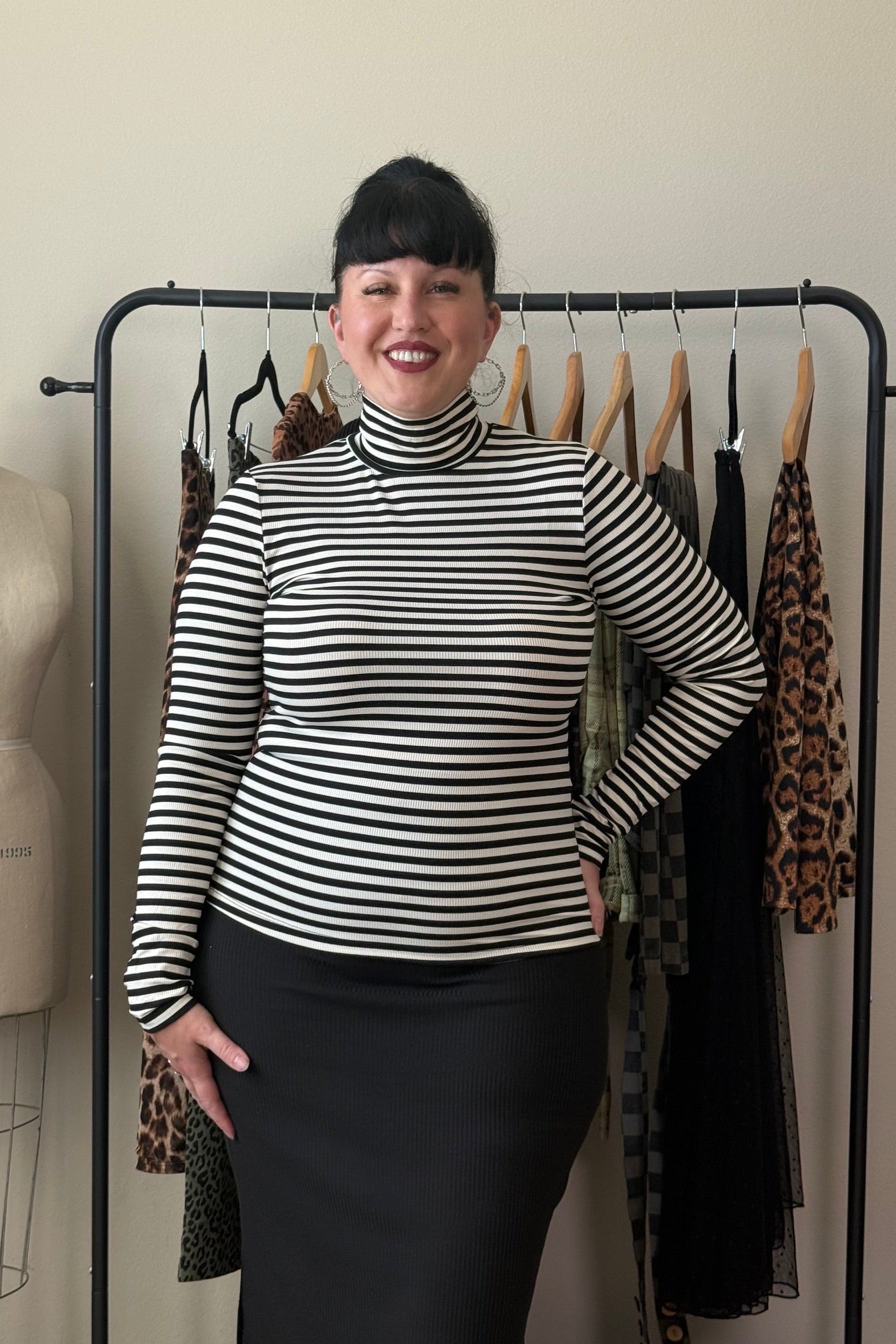 A woman with short black hair stands smiling in front of a clothing rack, wearing a black and white horizontally striped mock neck long-sleeve top and a black skirt. A dress form is visible to her left.