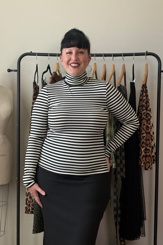 A woman with short black hair stands smiling in front of a clothing rack, wearing a black and white horizontally striped mock neck long-sleeve top and a black skirt. A dress form is visible to her left.