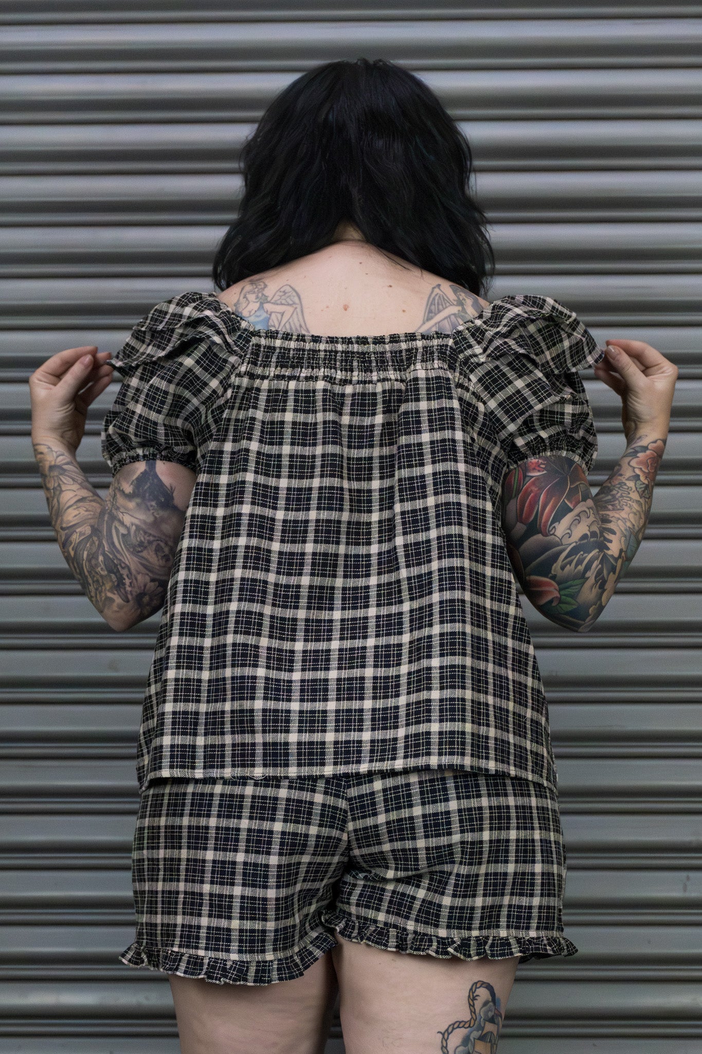 Person wearing a plaid outfit with tattoos visible, standing against a metallic shutter.