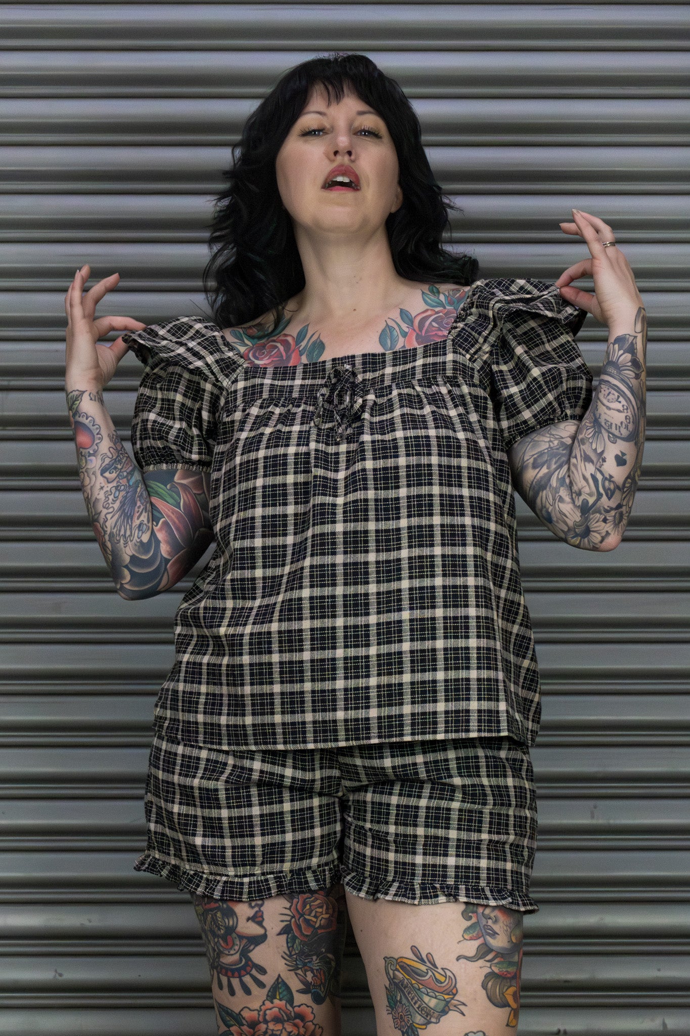 Person wearing a plaid outfit with tattoos standing against a metallic shutter.