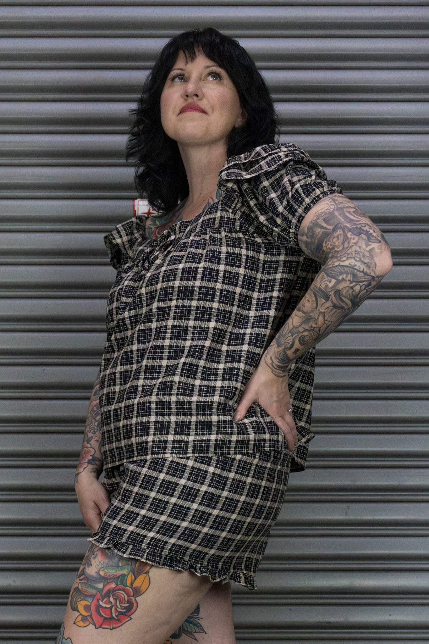 Person wearing a plaid dress with tattoos standing against a metallic shutter.