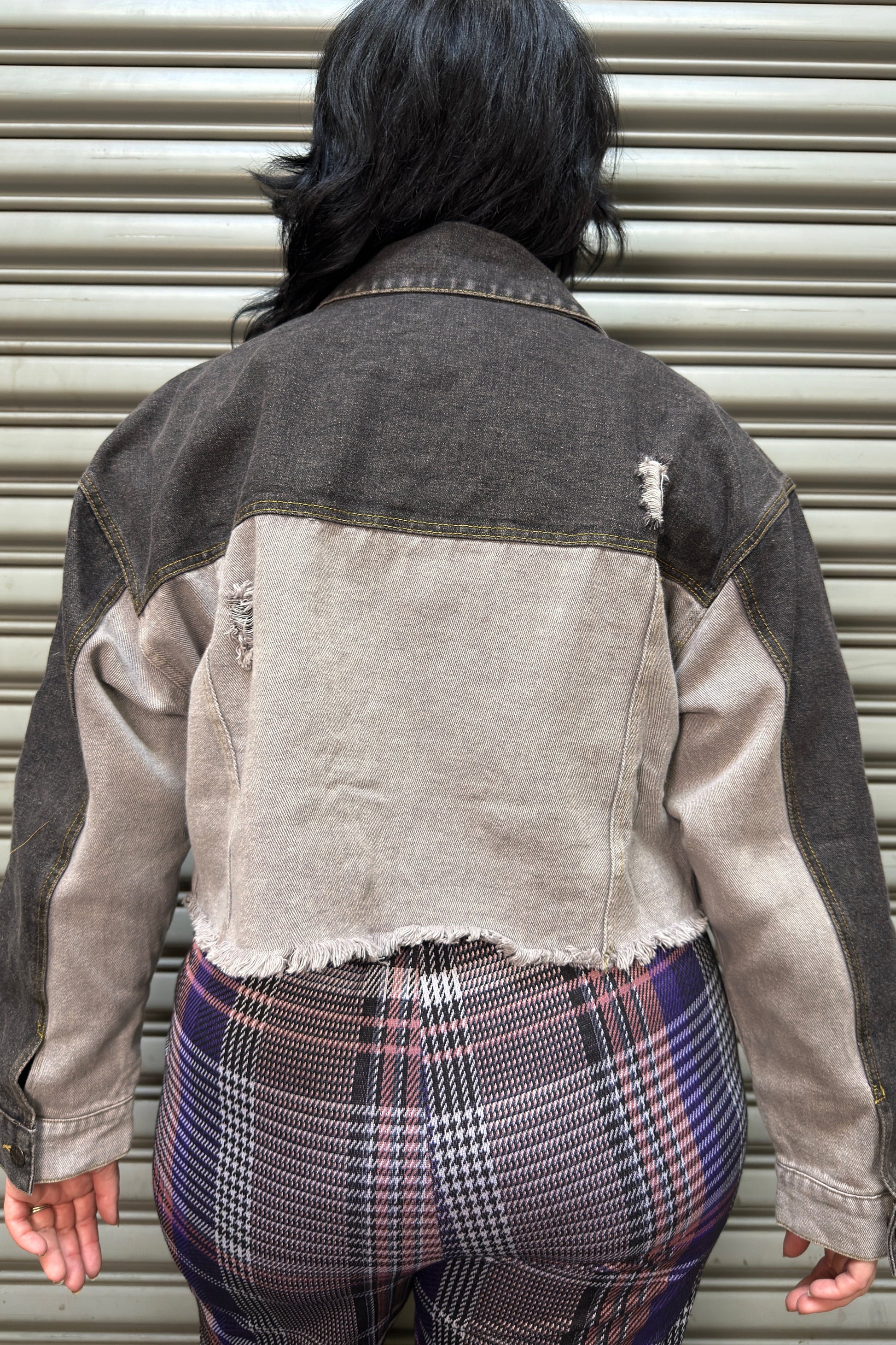 Person wearing a denim jacket and plaid pants standing in front of a metal shutter.