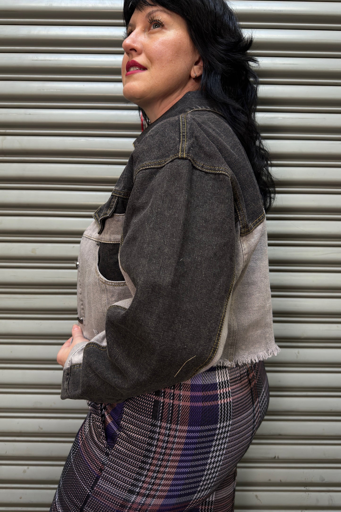 Woman wearing a denim jacket and plaid skirt standing against a metal shutter.