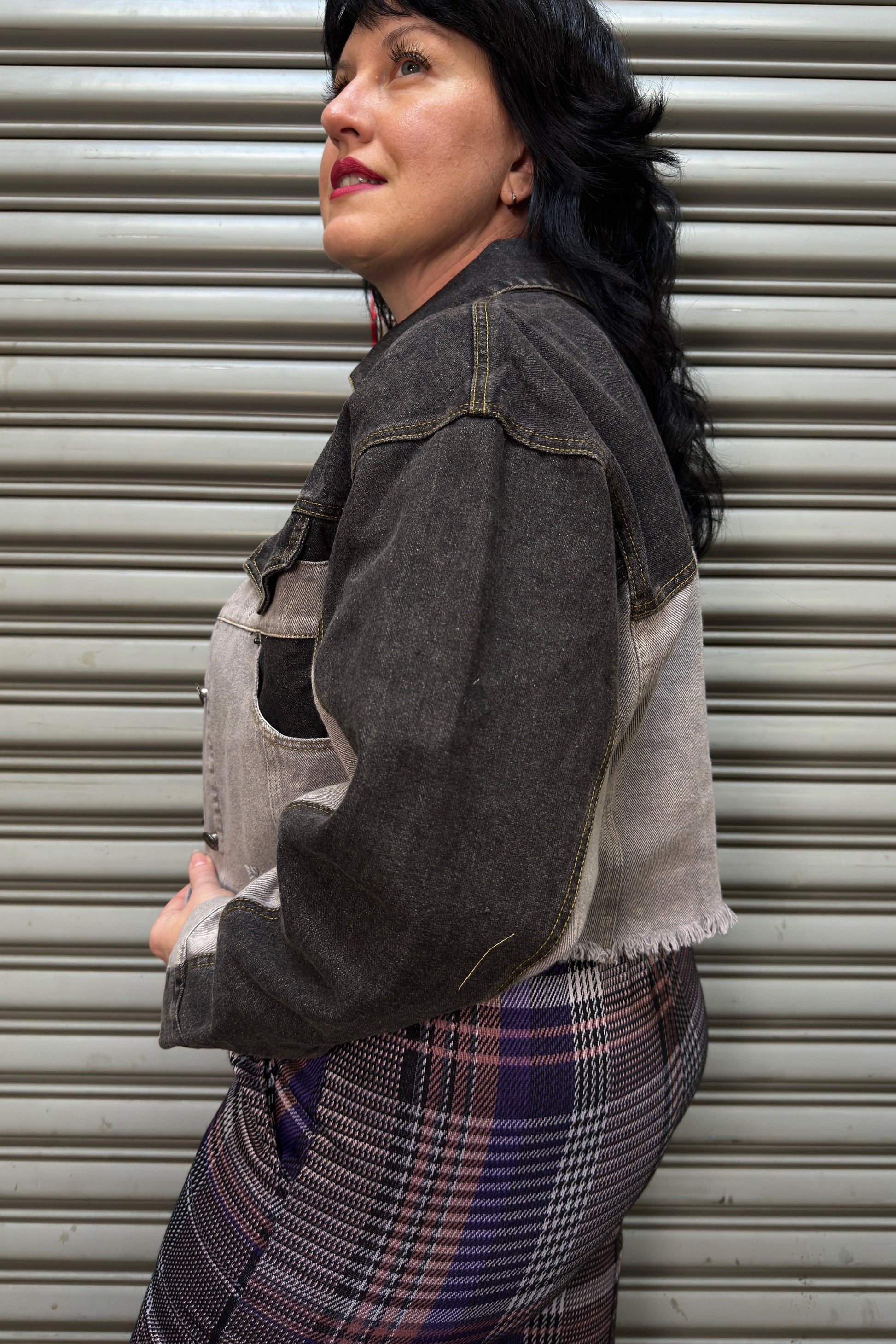 Woman wearing a denim jacket and plaid skirt standing against a metal shutter.