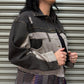 Woman wearing a dark denim jacket with a gray patch over a patterned outfit, standing against a metallic shutter.