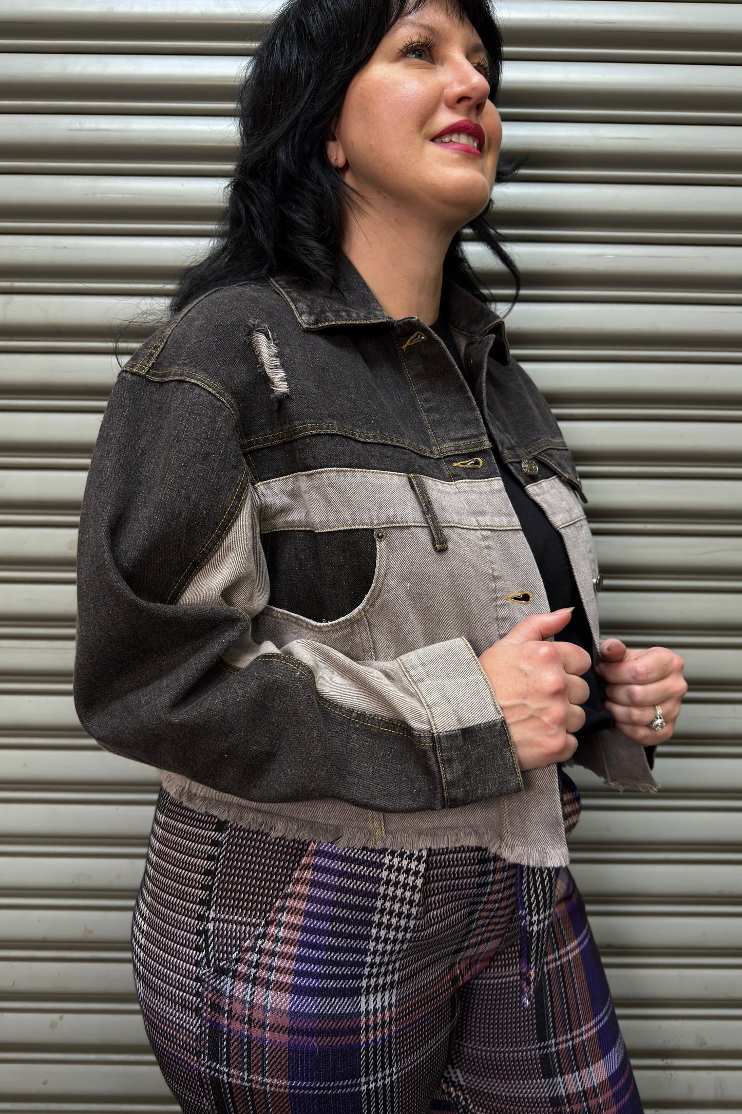 Woman wearing a dark denim jacket with a gray patch over a patterned outfit, standing against a metallic shutter.