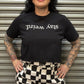 Person wearing a black t-shirt with 'stay weird' text and checkered pants in front of a metallic shutter.
