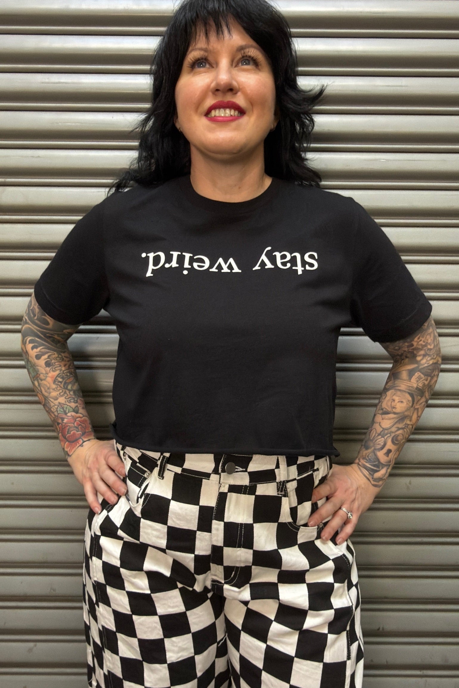 Person wearing a black t-shirt with 'stay weird' text and checkered pants in front of a metallic shutter.