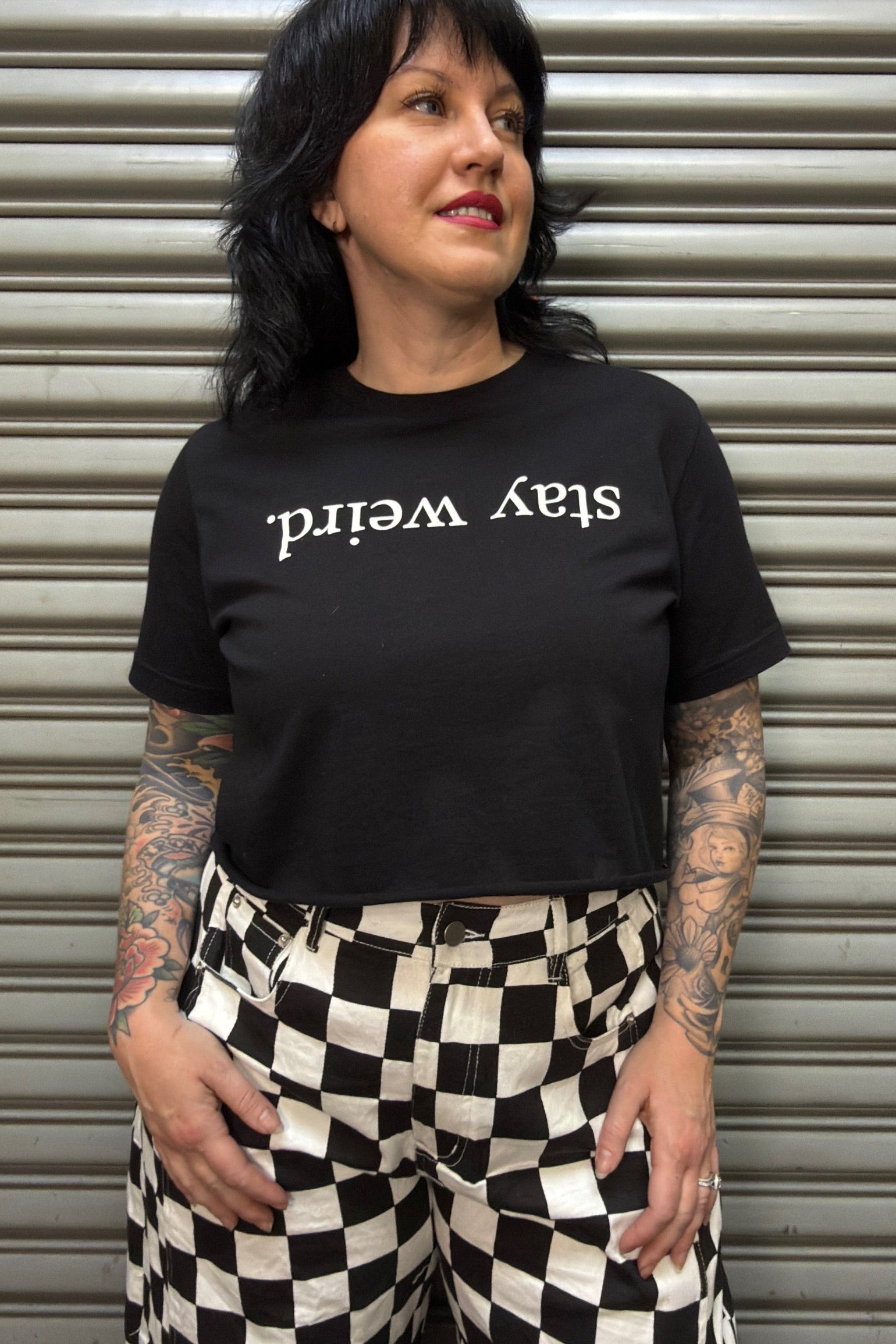 Person wearing a black t-shirt with 'stay weird' text in front of a metallic shutter.